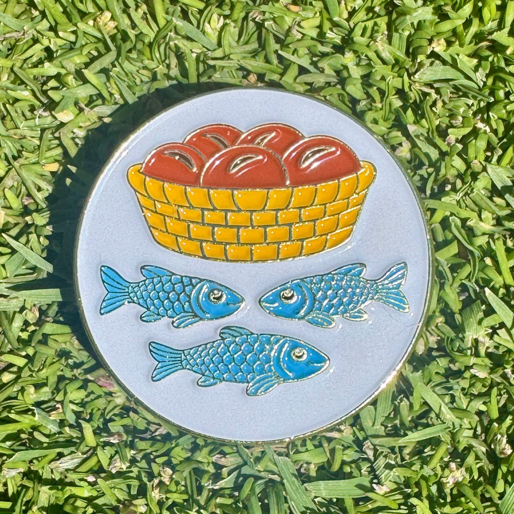 Metallic golf ball marker with a basket of apples and two blue fish on grass